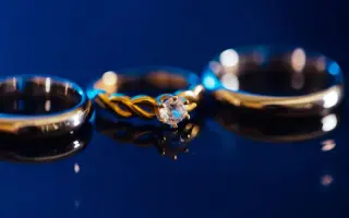 Two gold wedding bands and an engagement ring on a glossy surface against a deep blue background