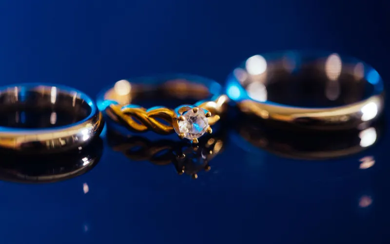 Two gold wedding bands and an engagement ring on a glossy surface against a deep blue background